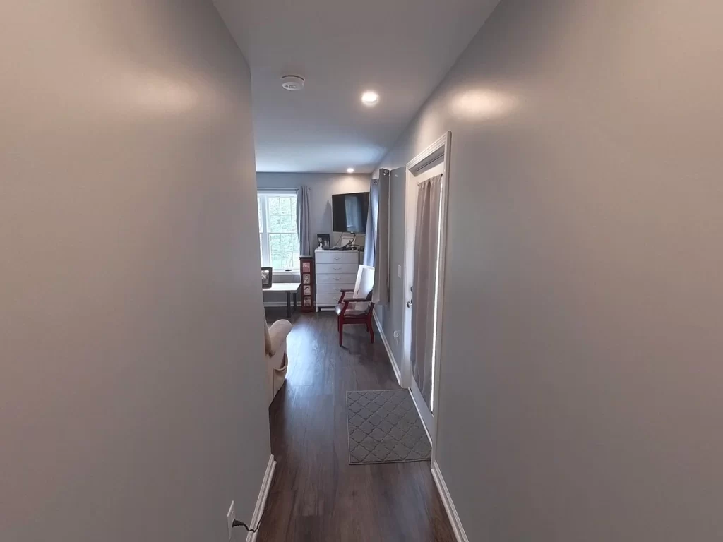 Hallway view leading into a newly remodeled in-law suite, featuring modern gray walls, wood flooring, and ample lighting, with a glimpse of a comfortable living space including a recliner, bed, flat-screen TV, and dresser.