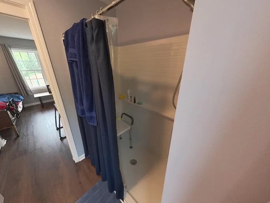 Accessible bathroom in a newly remodeled in-law suite, featuring a walk-in shower with grab bars, blue towels and bath mat, gray curtains, and modern fixtures designed for elder family member care.