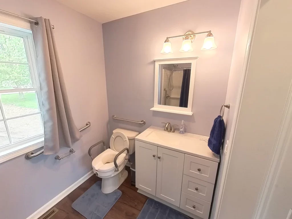 Accessible bathroom in a newly remodeled in-law suite, featuring a toilet with grab bars, a sink with a mirror and vanity, and a view into the adjacent bedroom. The space has modern gray walls, wood flooring, and ample lighting, designed for elder family member care.