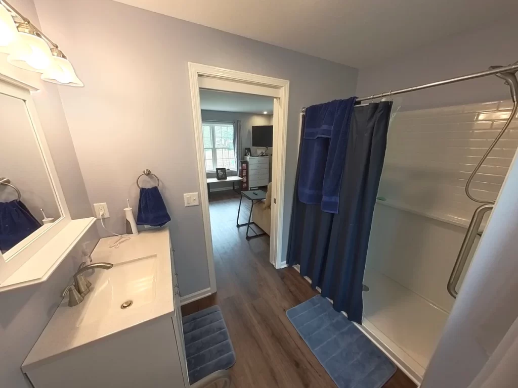 Accessible bathroom in a newly remodeled in-law suite, featuring a sink with vanity and mirror, walk-in shower with grab bars, and blue towels. The bathroom opens into a bedroom with modern gray walls, wood flooring, and ample lighting, designed for elder family member care.