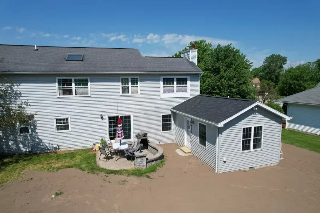 Exterior view of a completed home addition, featuring a new in-law suite with gray siding and a black roof, connected to the main house. The backyard includes a patio area with outdoor furniture and a grill, set against a clear blue sky.