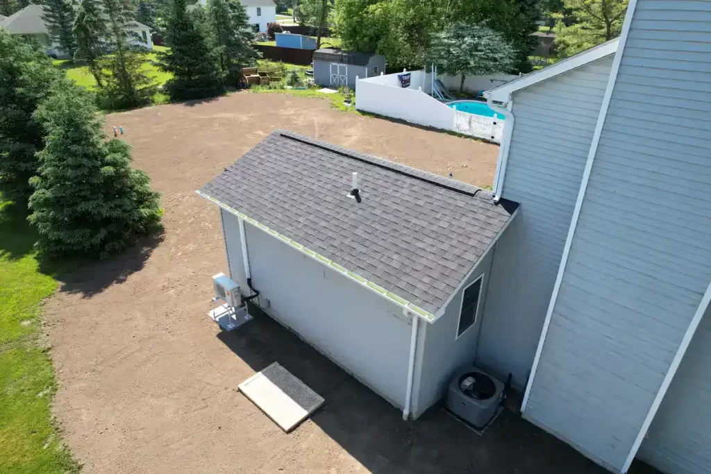 Aerial view of the side of a completed home addition, featuring gray siding and a black roof, connected to the main house. The backyard is spacious with freshly leveled soil, surrounded by trees and neighboring homes.