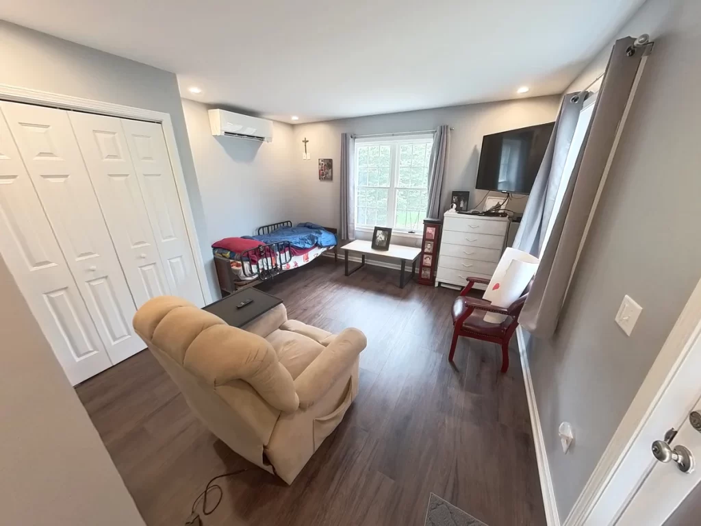 Interior of a newly remodeled in-law suite, featuring a comfortable recliner, bed, flat-screen TV, dresser, and large window, with modern gray walls, wood flooring, and ample lighting, designed for elder family member care.