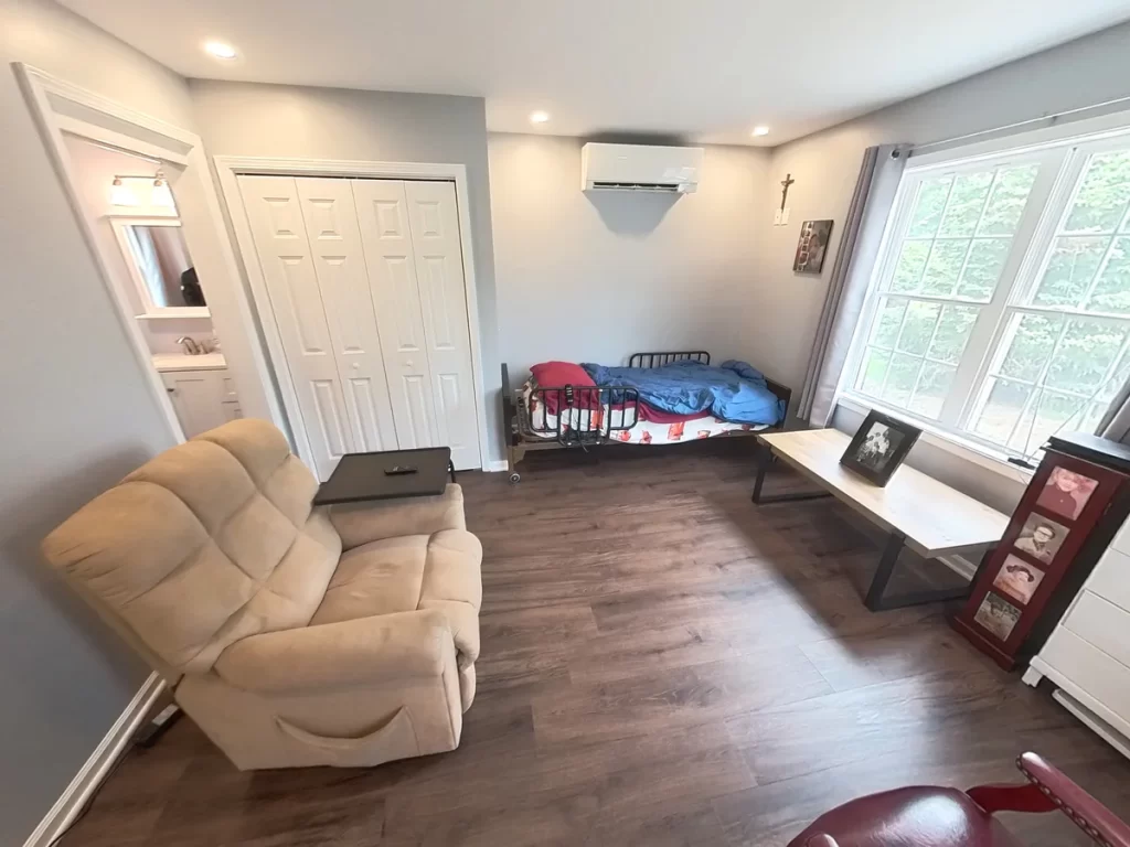 Interior of a newly remodeled in-law suite, featuring a comfortable recliner, bed, flat-screen TV, dresser, and large window, with modern gray walls, wood flooring, and ample lighting, designed for elder family member care.