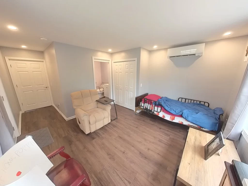 Interior of a newly remodeled in-law suite, featuring a comfortable recliner, bed, flat-screen TV, dresser, and large window, with modern gray walls, wood flooring, and ample lighting, designed for elder family member care.