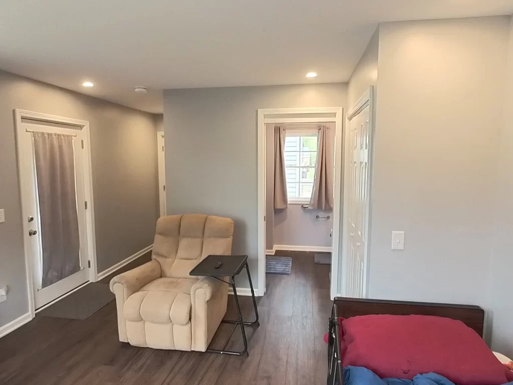 Interior of a newly remodeled in-law suite, featuring a beige recliner, a bed, and a hallway leading to a bathroom. The space has modern gray walls, wood flooring, and ample lighting, designed for elder family member care.