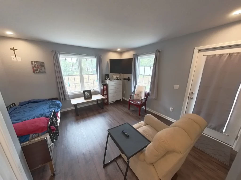 Interior of a newly remodeled in-law suite, featuring a comfortable recliner, bed, flat-screen TV, dresser, and large window, with modern gray walls, wood flooring, and ample lighting, designed for elder family member care.