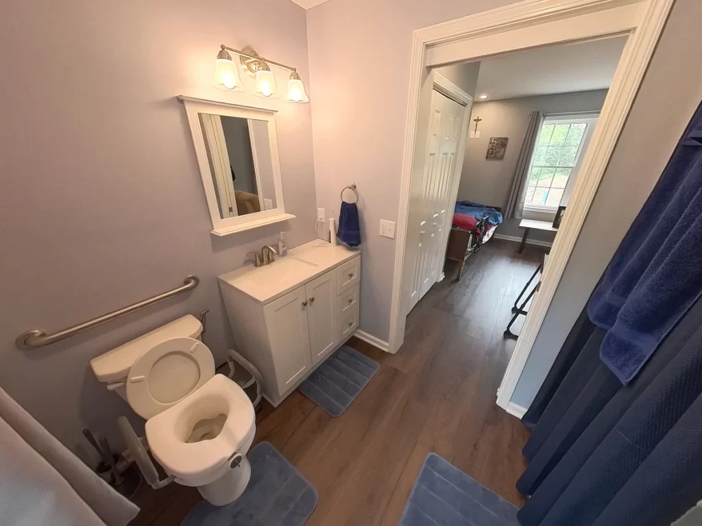 Accessible bathroom in a newly remodeled in-law suite, featuring a toilet with grab bars, a sink with a mirror and vanity, and a view into the adjacent bedroom. The space has modern gray walls, wood flooring, and ample lighting, designed for elder family member care.