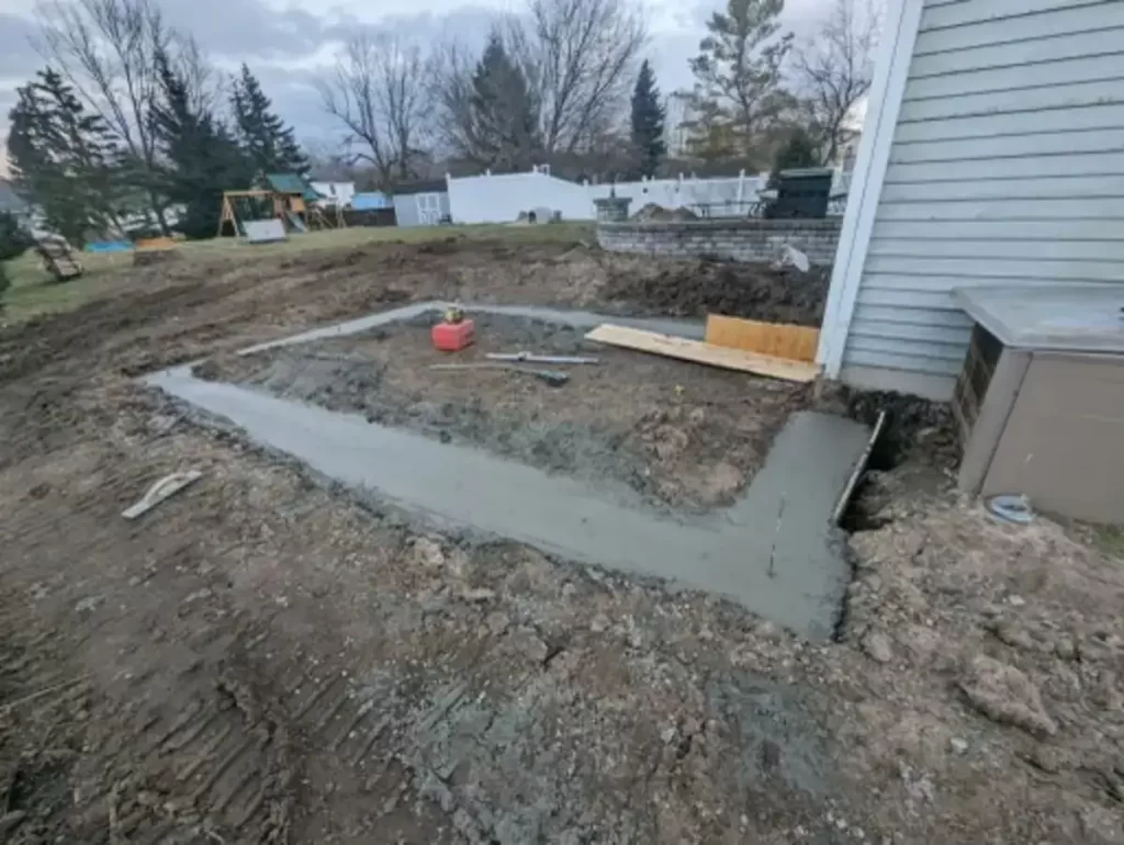 Foundation being laid for a home addition, showing early construction stages with fresh concrete, adjacent to an existing house structure, in a residential backyard setting.