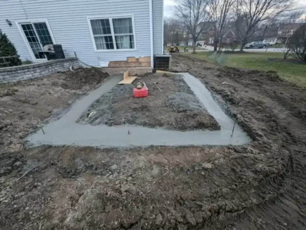 Foundation being laid for a home addition, showing early construction stages with fresh concrete, adjacent to an existing house structure, in a residential backyard setting.