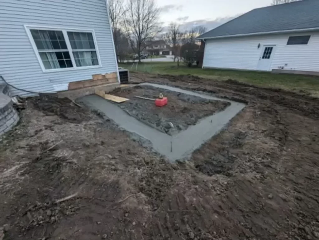 Foundation being laid for a home addition, showing early construction stages with fresh concrete, adjacent to an existing house structure, in a residential backyard setting.