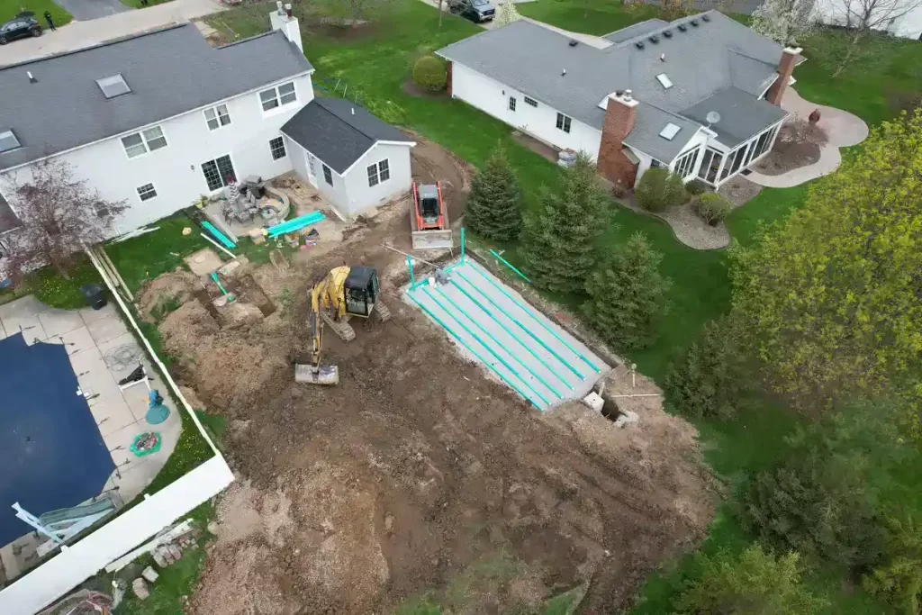 Aerial view of a septic system installation for a home addition project, featuring excavation machinery, PVC pipes, and a drain field setup adjacent to an existing house in a suburban backyard.