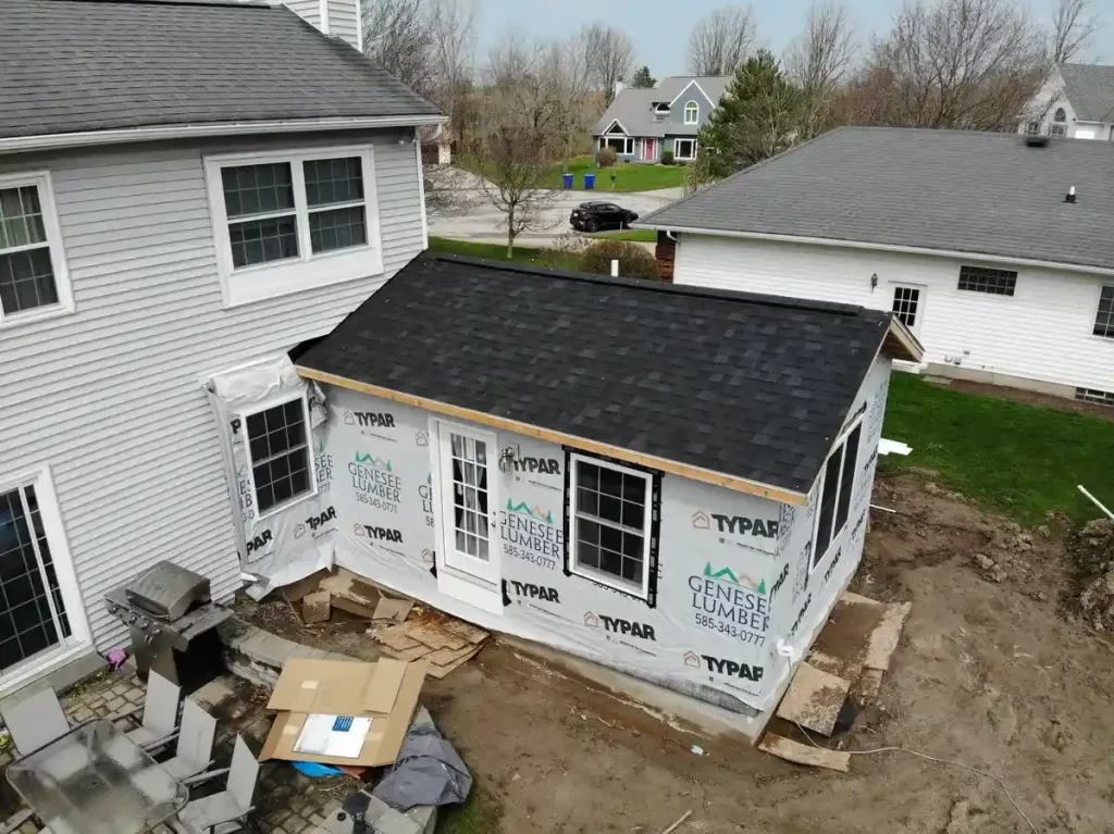 Partially completed home addition under construction, with new windows, doors, and roofing, covered in Typar and Genesee Lumber wrap, adjacent to an existing house in a suburban neighborhood.