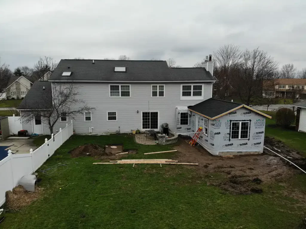 Partially completed home addition under construction, with new windows, doors, and roofing, covered in Typar and Genesee Lumber wrap, adjacent to an existing house in a suburban neighborhood.