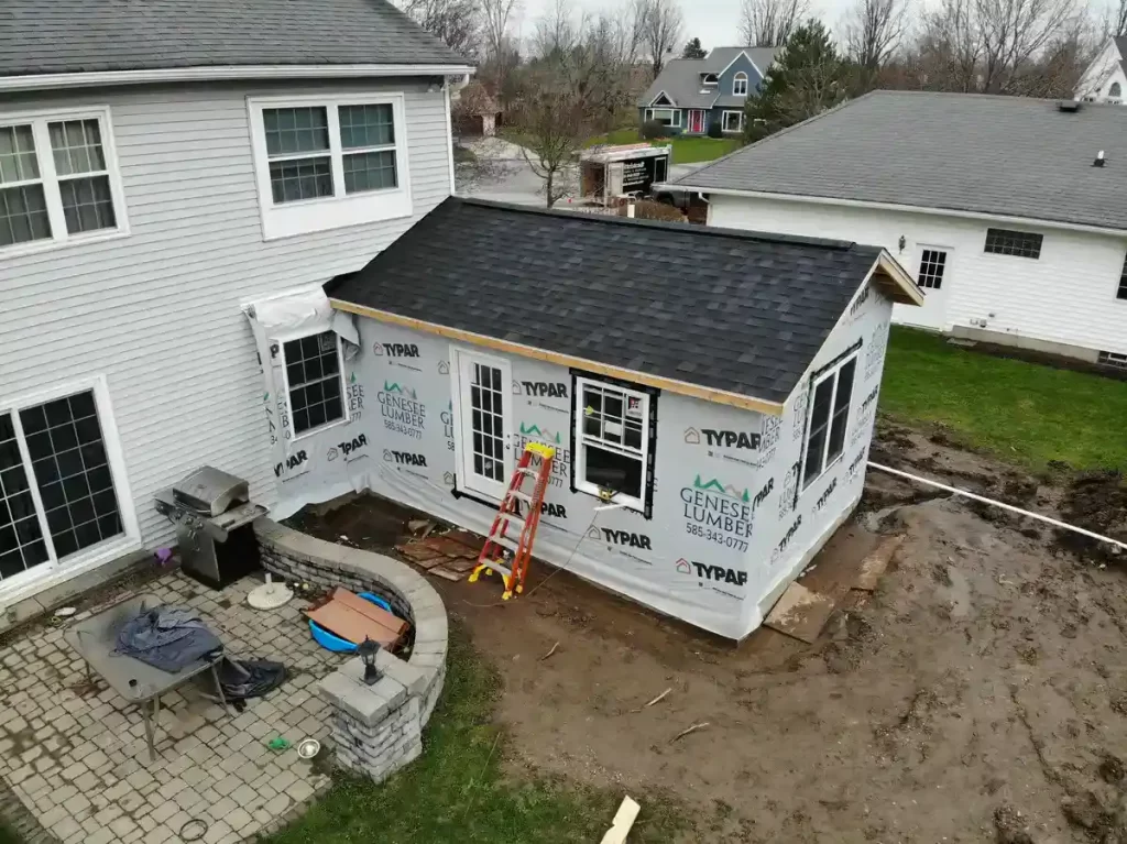 Partially completed home addition under construction, with new windows, doors, and roofing, covered in Typar and Genesee Lumber wrap, adjacent to an existing house in a suburban neighborhood.