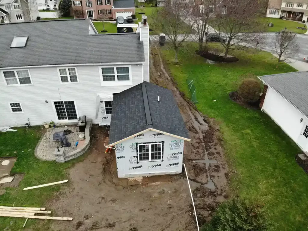 Partially completed home addition under construction, with new windows, doors, and roofing, covered in Typar and Genesee Lumber wrap, adjacent to an existing house in a suburban neighborhood.