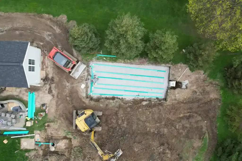 Aerial view of a septic system installation for a home addition project, featuring excavation machinery, PVC pipes, and a drain field setup adjacent to an existing house in a suburban backyard.