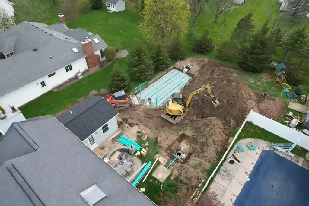 Aerial view of a septic system installation for a home addition project, featuring excavation machinery, PVC pipes, and a drain field setup adjacent to an existing house in a suburban backyard.