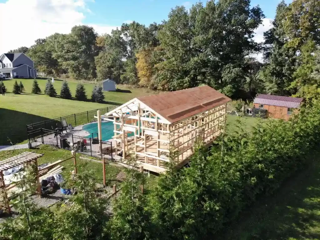 Aerial view of a pool house construction project with the frame structure completed, situated beside a fenced pool and lush green yard.