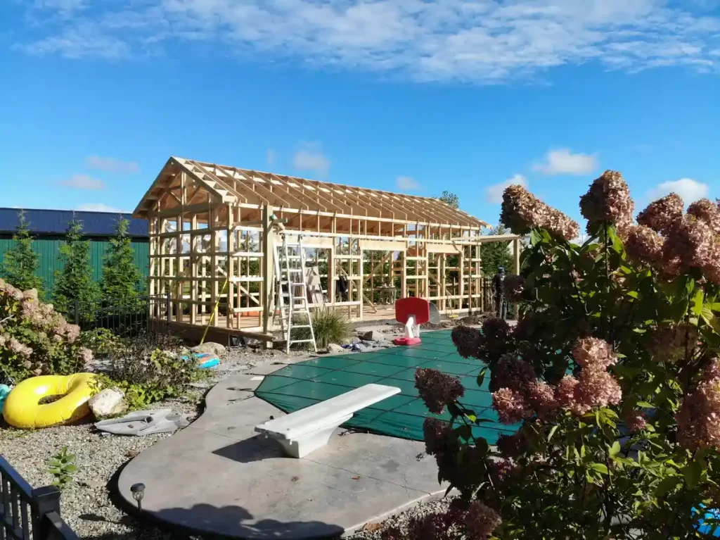 Ground-level view of the pool house frame being constructed next to a pool, with clear blue sky in the background.