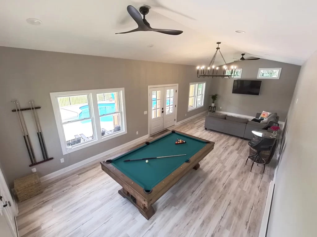 Interior view of the pool house addition in Akron, NY, showcasing the pool table, sectional sofa, and windows opening to the pool area, designed by Stately General Contracting.