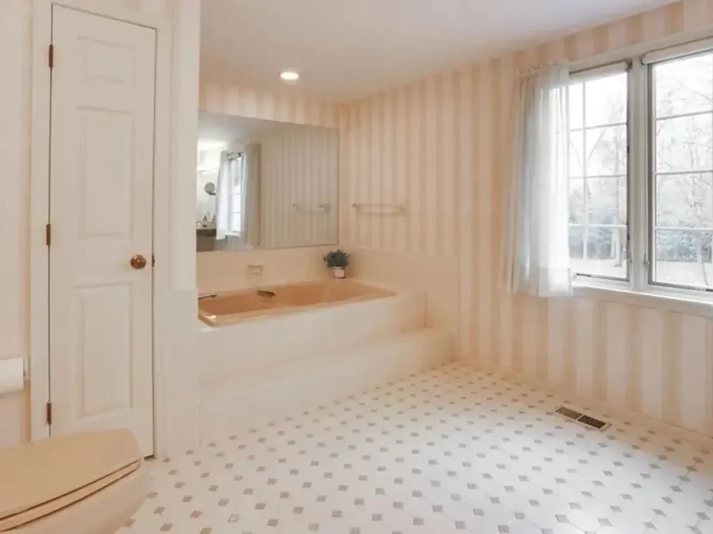 Before photo of an outdated bathroom featuring a built-in tub with beige fixtures, striped wallpaper, large window, and tile flooring.