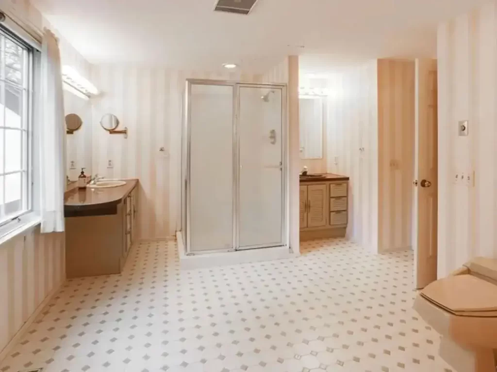 Before photo of an outdated bathroom showing a shower stall, double vanity, striped wallpaper, and hexagonal tile flooring with beige tones.