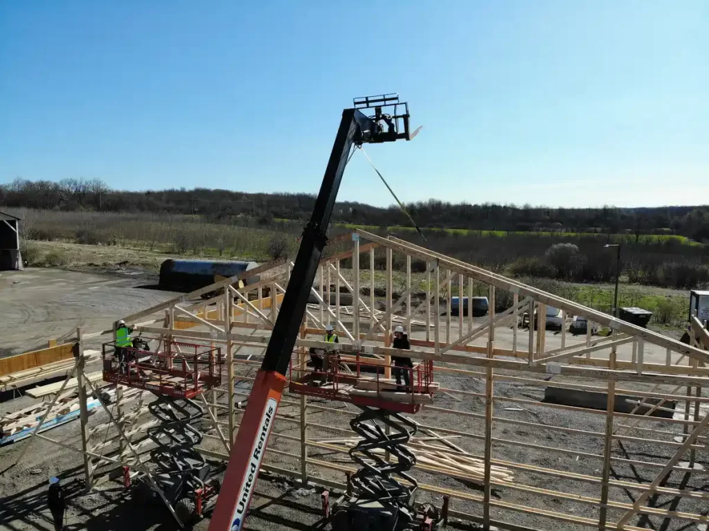 Close-up view of construction workers assembling the frame of the Bennington Highway Department pole barn using scissor lifts and heavy machinery to secure the trusses in place.