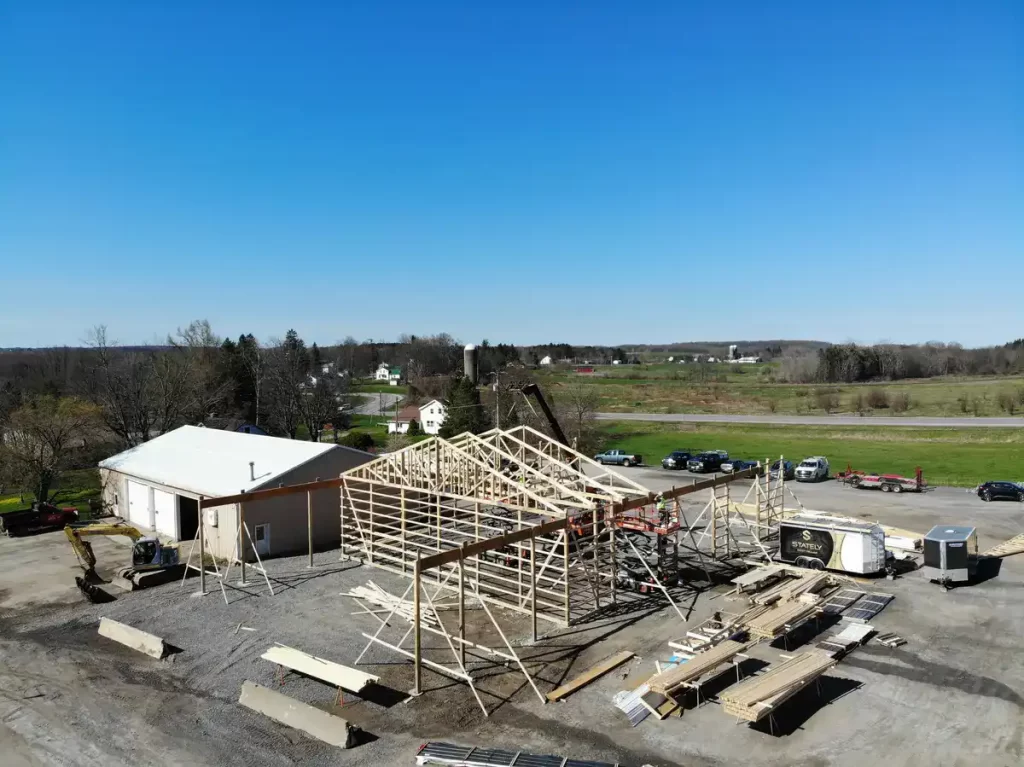 Aerial view of the Bennington Highway Department pole barn construction during the framing stage, showing the progress of the roof trusses being installed and construction materials organized on-site.