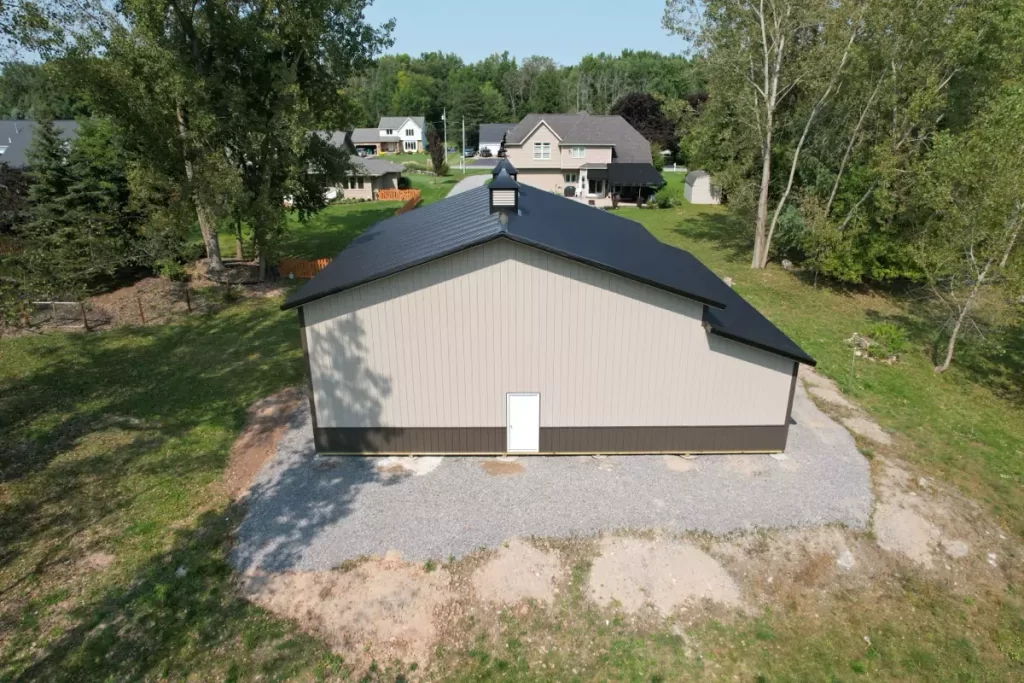 Rear view of a finished Stately Pole Barn showing the black metal roof, gravel foundation, and surrounding trees.