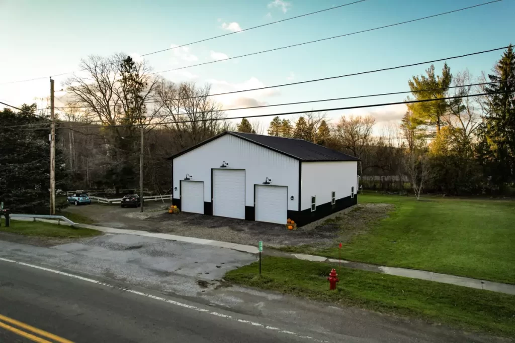 Durable white and black post-frame structure by Stately Pole Barns in Holland, NY, featuring three garage doors and a gravel driveway.