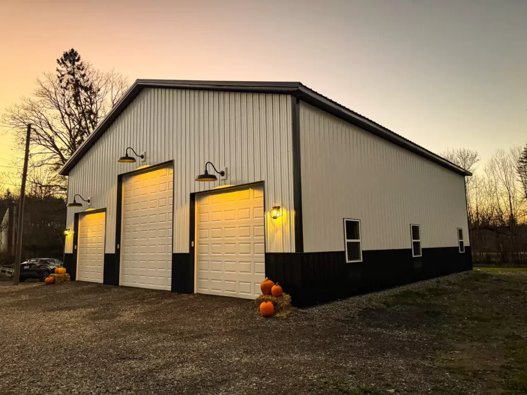 Stately Pole Barn with lit garage doors and pumpkins at twilight, showcasing aesthetic lighting and custom craftsmanship in Holland, NY.