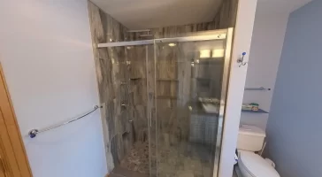 After renovation, an inviting walk-in shower with clear glass door, modern gray tile design, and high-quality fixtures is the centerpiece of this bathroom, highlighting the expertise of Stately Kitchen and Bath in delivering stylish and practical bathroom transformations.