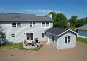 Exterior view of a completed home addition, featuring a new in-law suite with gray siding and a black roof, connected to the main house. The backyard includes a patio area with outdoor furniture and a grill, set against a clear blue sky.