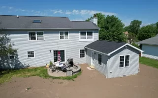 Exterior view of a completed home addition, featuring a new in-law suite with gray siding and a black roof, connected to the main house. The backyard includes a patio area with outdoor furniture and a grill, set against a clear blue sky.