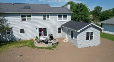 Exterior view of a completed home addition, featuring a new in-law suite with gray siding and a black roof, connected to the main house. The backyard includes a patio area with outdoor furniture and a grill, set against a clear blue sky.