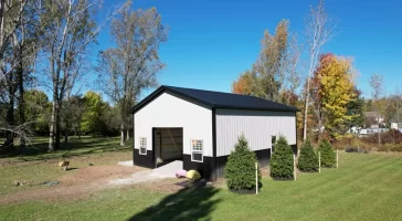 Side view of Stately Pole Barn with black wainscot, surrounded by greenery and trees.