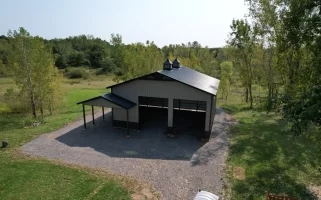 Aerial view of a finished Stately Pole Barn with black roofing, two cupolas, and surrounding green landscape.