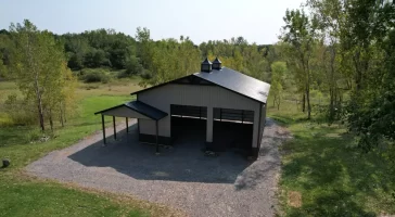 Aerial view of a finished Stately Pole Barn with black roofing, two cupolas, and surrounding green landscape.