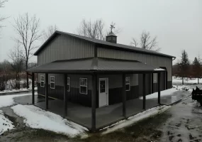 Frontal view of a pole barn with a centered man door, flanked by windows and garage doors, under a grey sky, with a thin layer of snow on the ground.