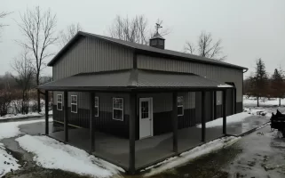 Frontal view of a pole barn with a centered man door, flanked by windows and garage doors, under a grey sky, with a thin layer of snow on the ground.