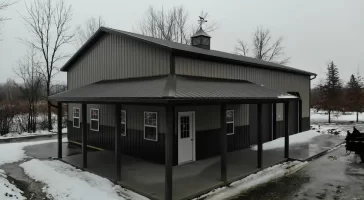 Frontal view of a pole barn with a centered man door, flanked by windows and garage doors, under a grey sky, with a thin layer of snow on the ground.